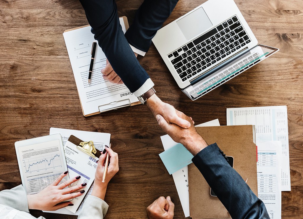 Overhead view of two people shaking hands over a desk with printed financial charts, documents and a laptop.