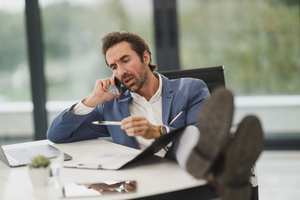 Man in a blue blazer leaning back with feet on a desk, talking on a phone and holding a pen