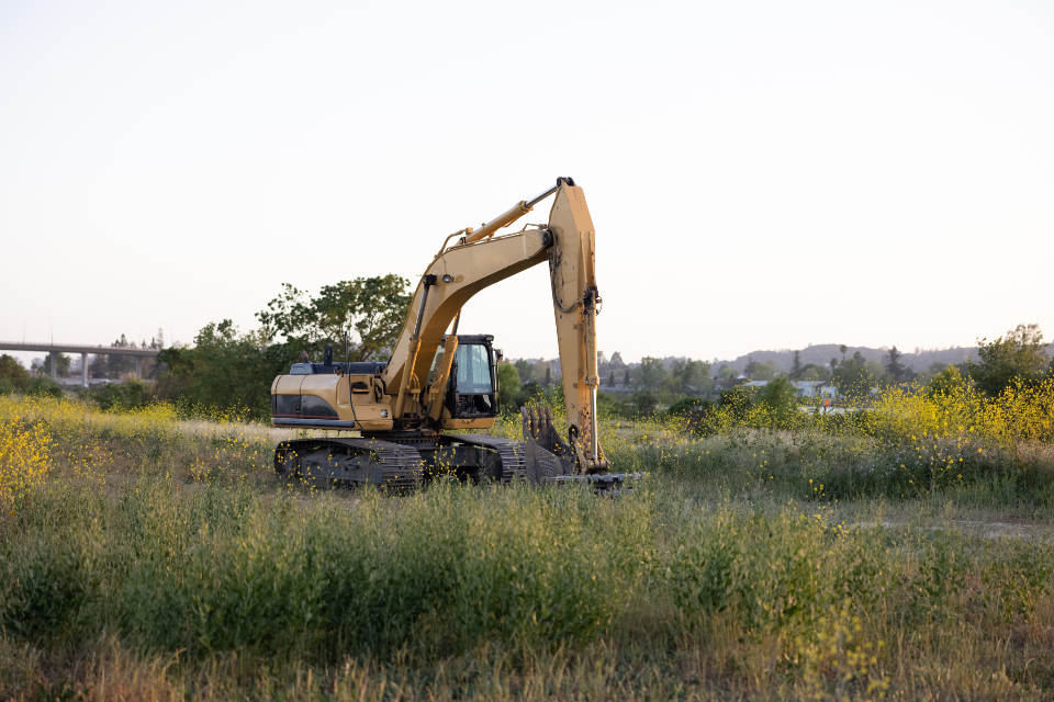 Yellow excavator with a long arm parked in tall grass and yellow wildflowers under a pale sky