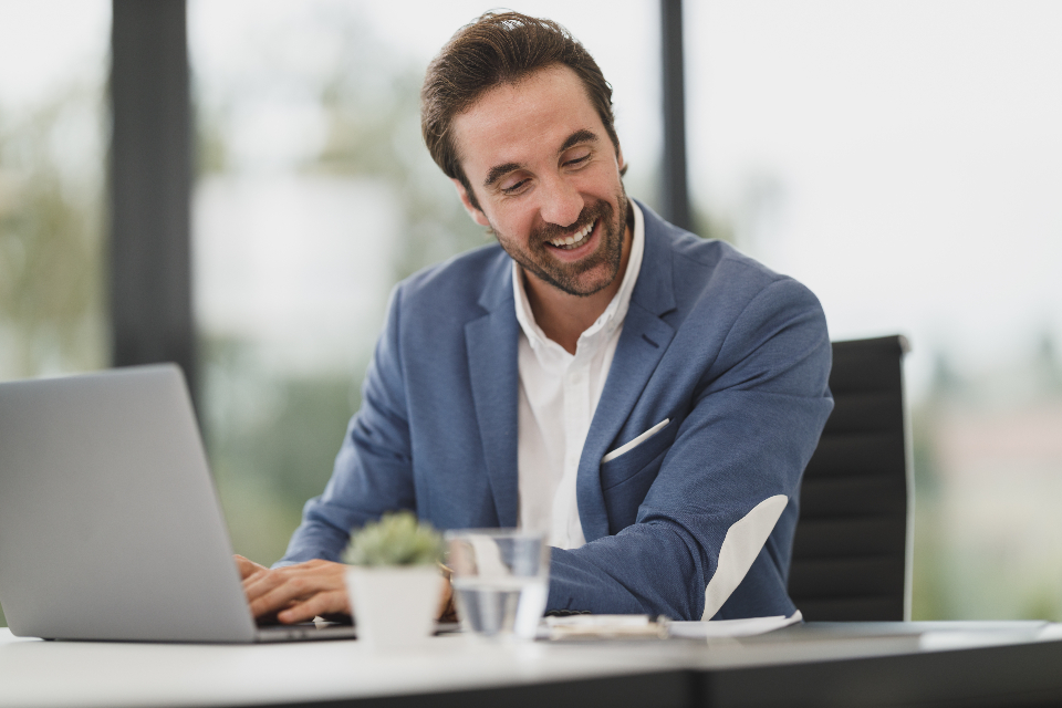 Smiling man in a blue blazer types on a laptop at a desk with a glass of water and a small potted plant.