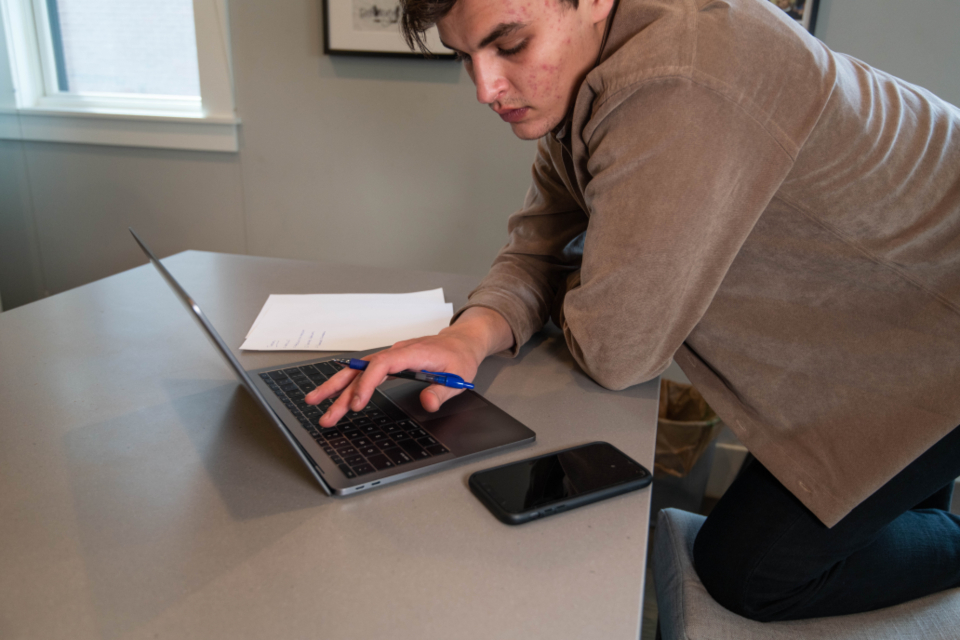 A young man leans over a laptop on a desk, holding a pen, with blank papers and a smartphone nearby.