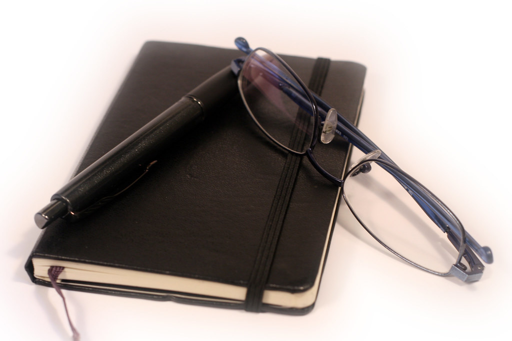 Black notebook with a black pen and blue-framed eyeglasses resting on top, on a white surface