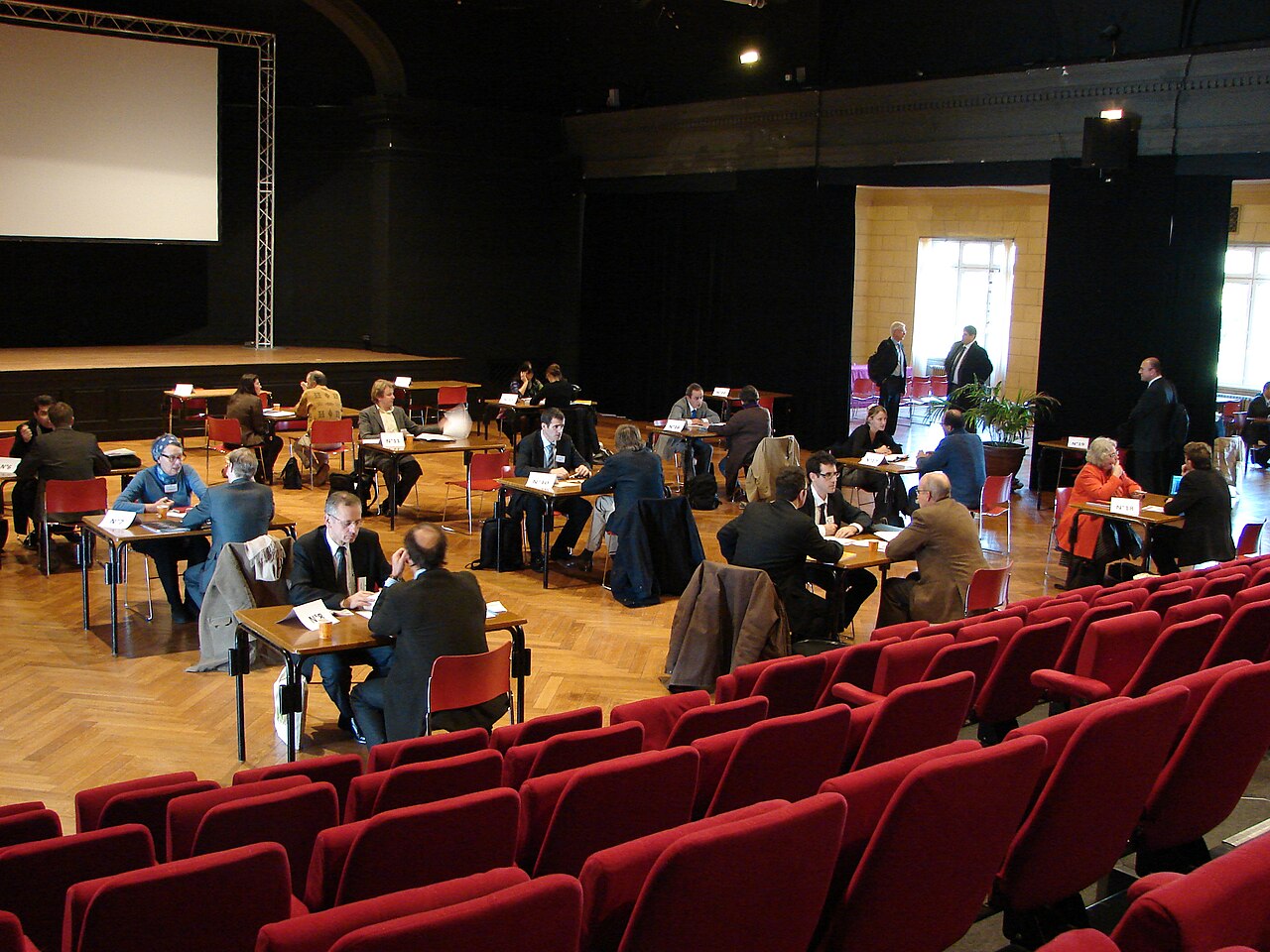 People seated at small tables with papers and name cards in a hall with a stage, screen and red seats