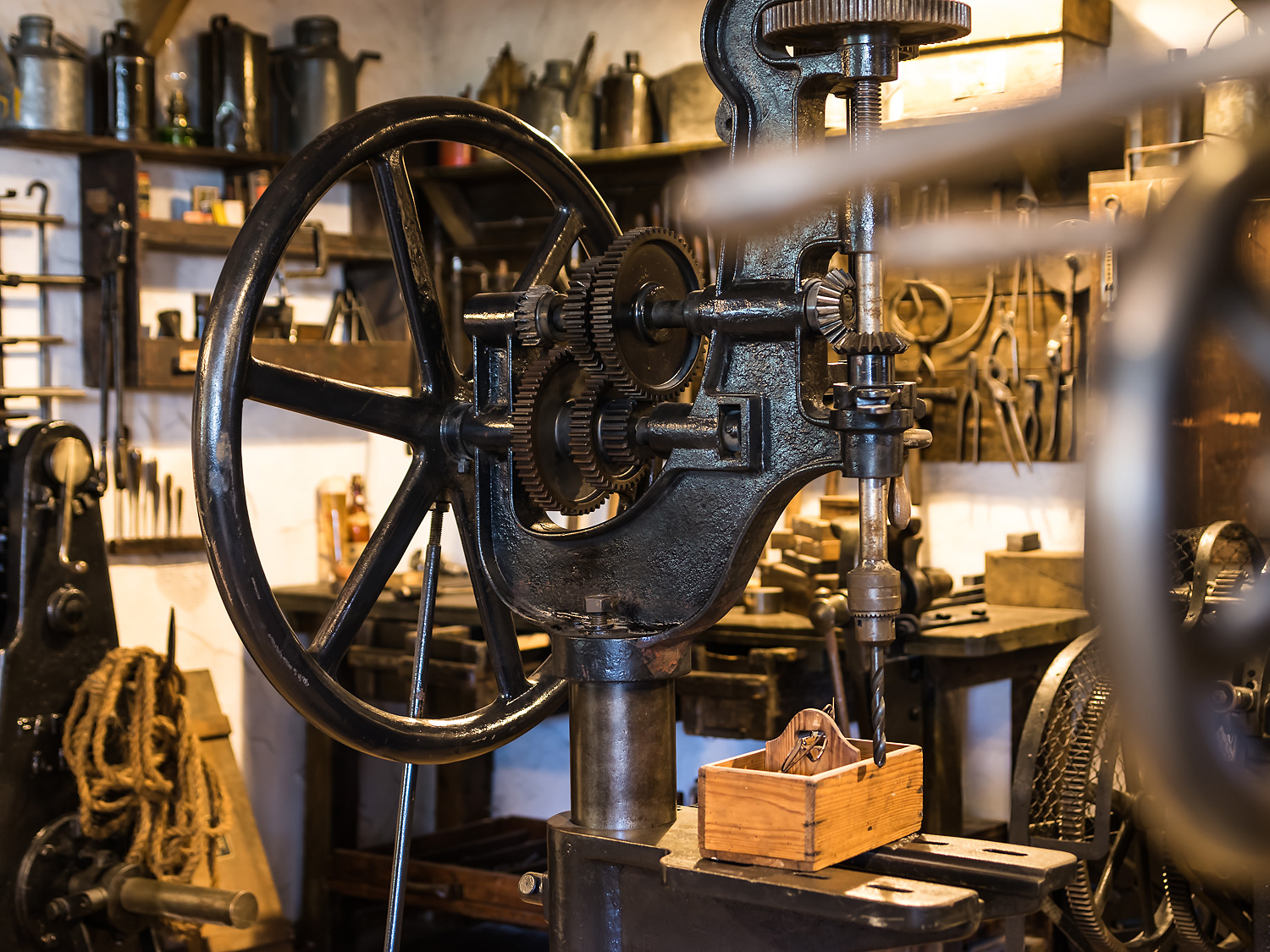 Antique black drill press with a large hand wheel in a cluttered workshop, tools on shelves behind.