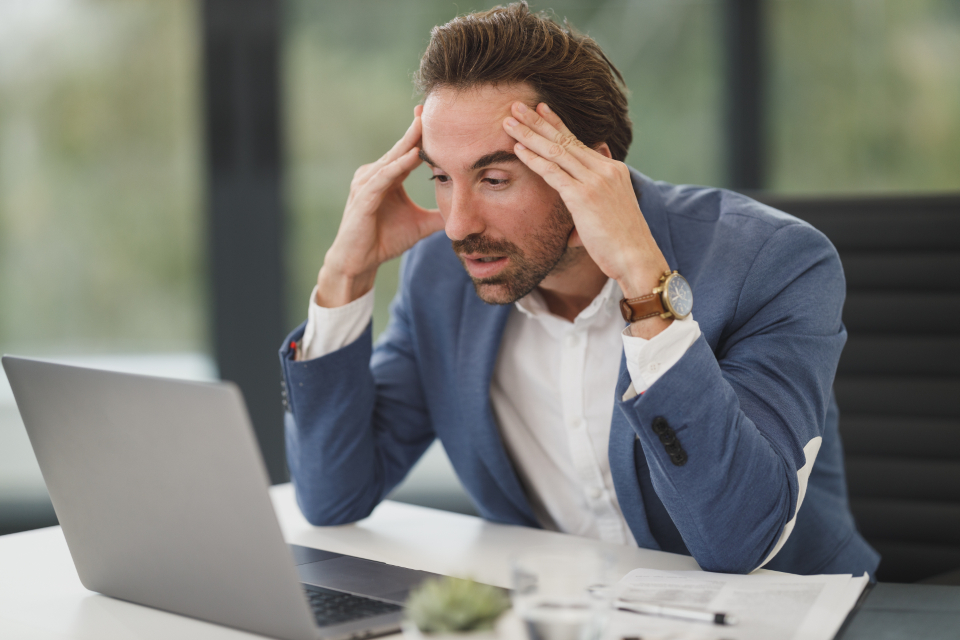 Stressed man in a blue blazer holding his head while sitting at a desk with a laptop and papers