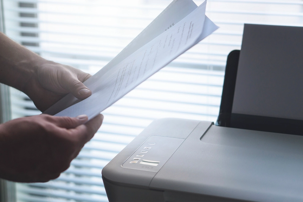 Hands holding a stack of printed white papers beside a printer in an office