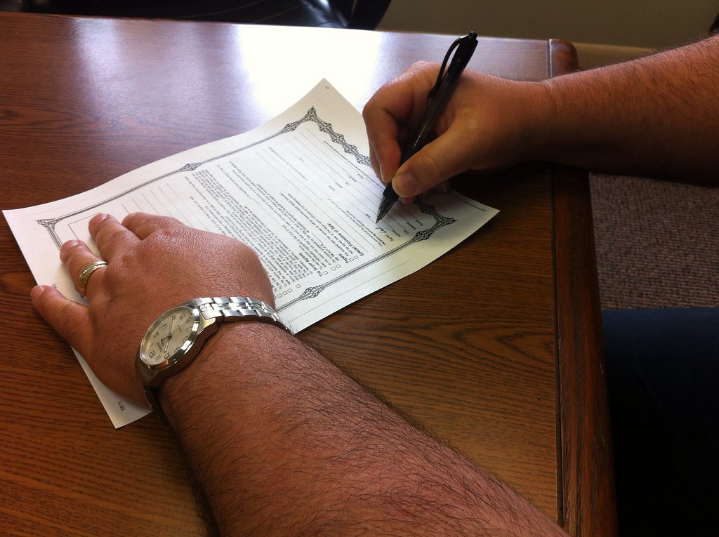 Close-up of a person's hands signing a printed document with a pen on a wooden desk, one hand wearing a watch and ring