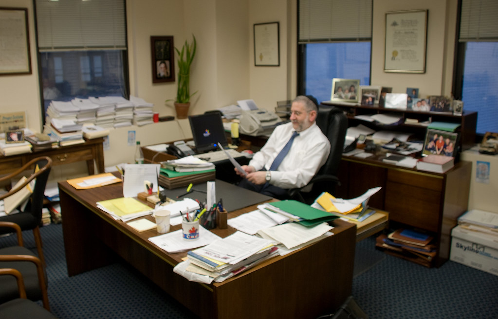 A man in a white shirt and tie sits at a cluttered wooden office desk reading printed papers