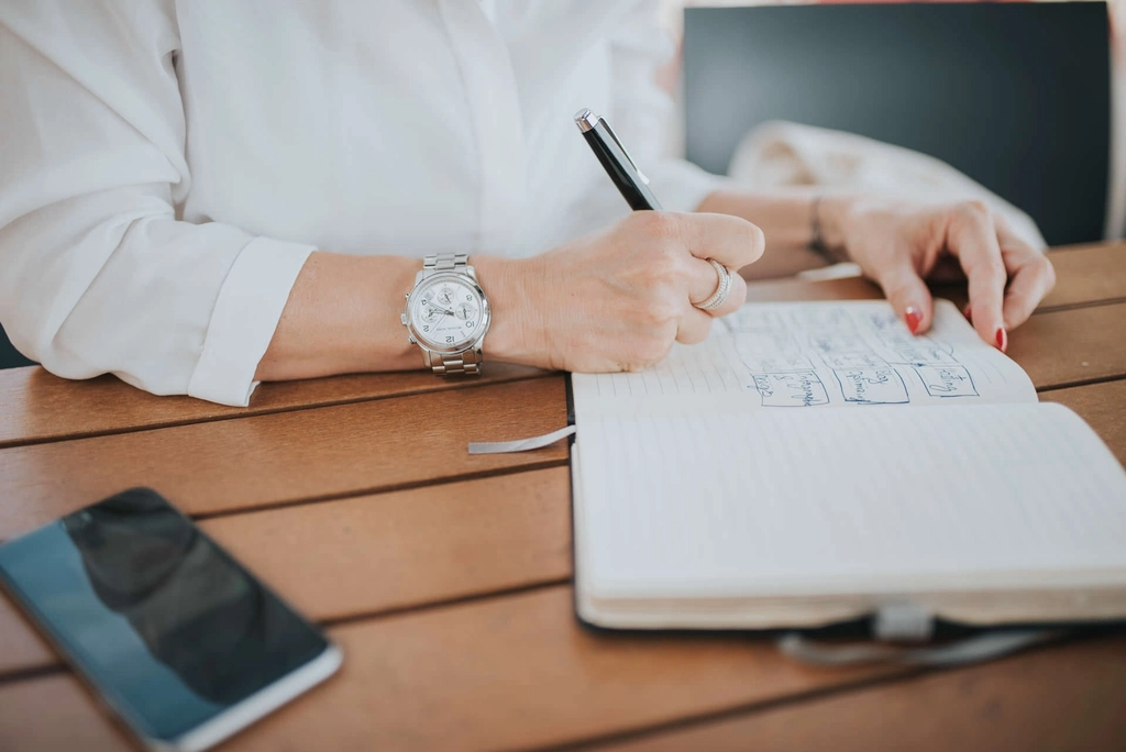 Person in a white shirt writing in a lined notebook with a pen, phone on a wooden table