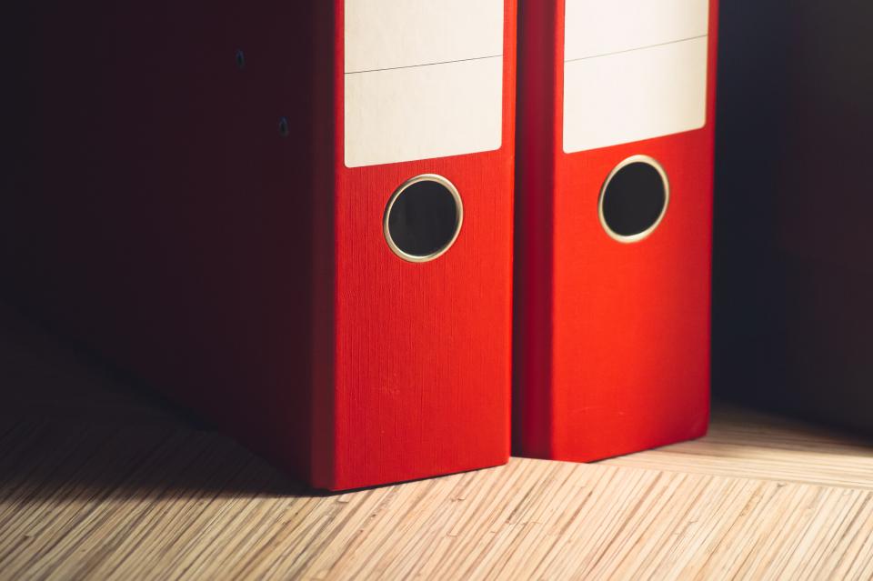 Two red ring binders standing side by side on a light wooden floor against a dark background