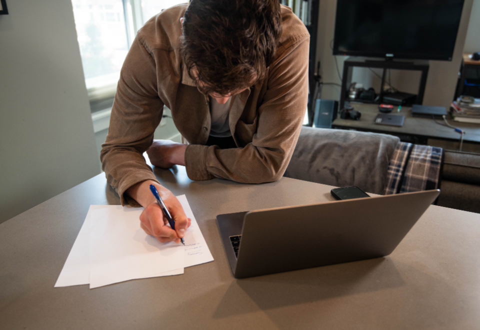 Person writing on blank sheets of paper at a table beside an open laptop