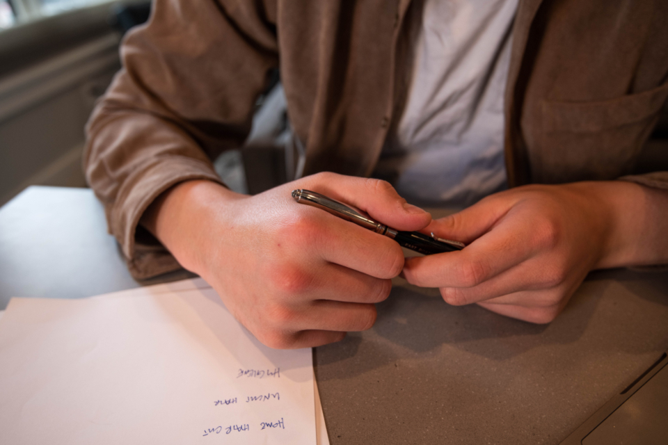 Close-up of hands holding a pen over a sheet of paper with handwritten notes on a grey desk.