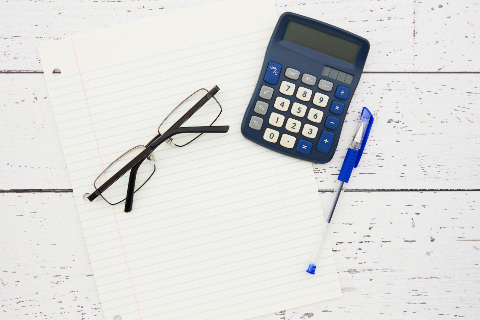 Lined notepad, black-framed eyeglasses, blue calculator and blue pen on a white wooden desk