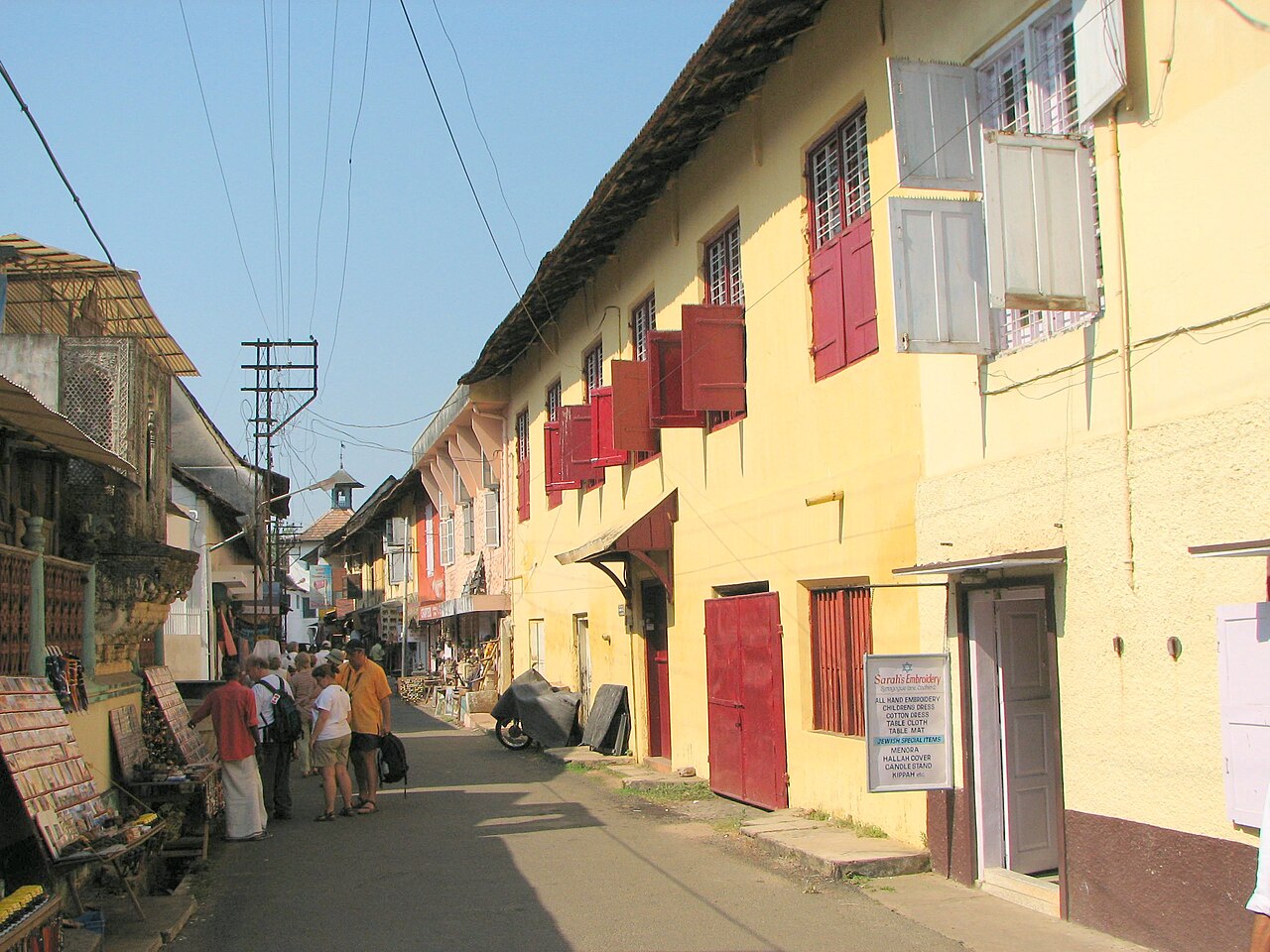 A sunlit street in an Indian market with yellow buildings, red shutters, a shop sign, and people walking.