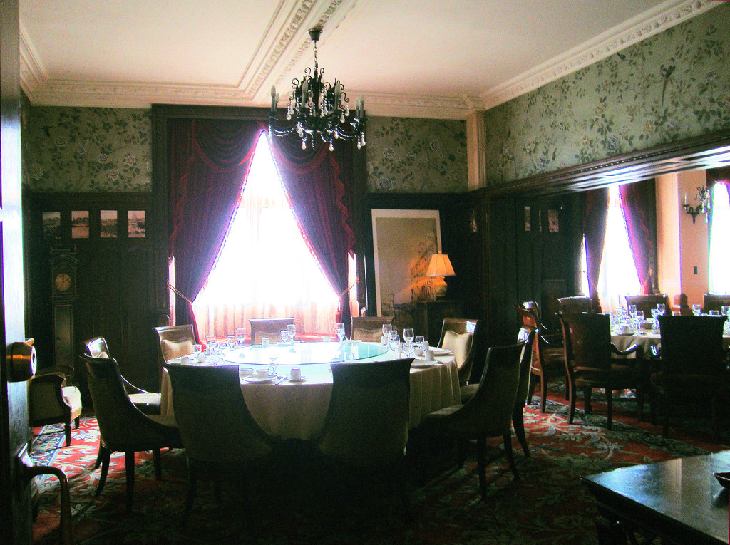 Formal dining room with a round table set with glasses and chairs, chandelier, floral wallpaper and red carpet.
