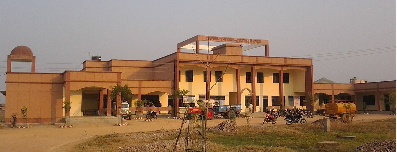 Government building with Hindi signage, arched entrance, parked motorcycles, a tractor-trolley and a water tanker