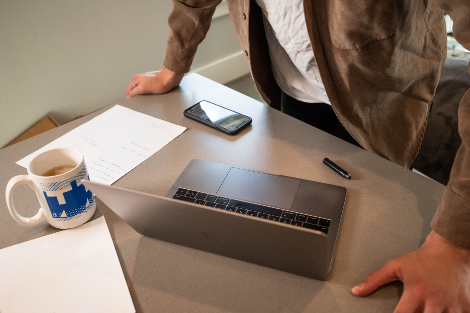 Person standing at a grey desk with an open laptop, phone, pen, mug and papers