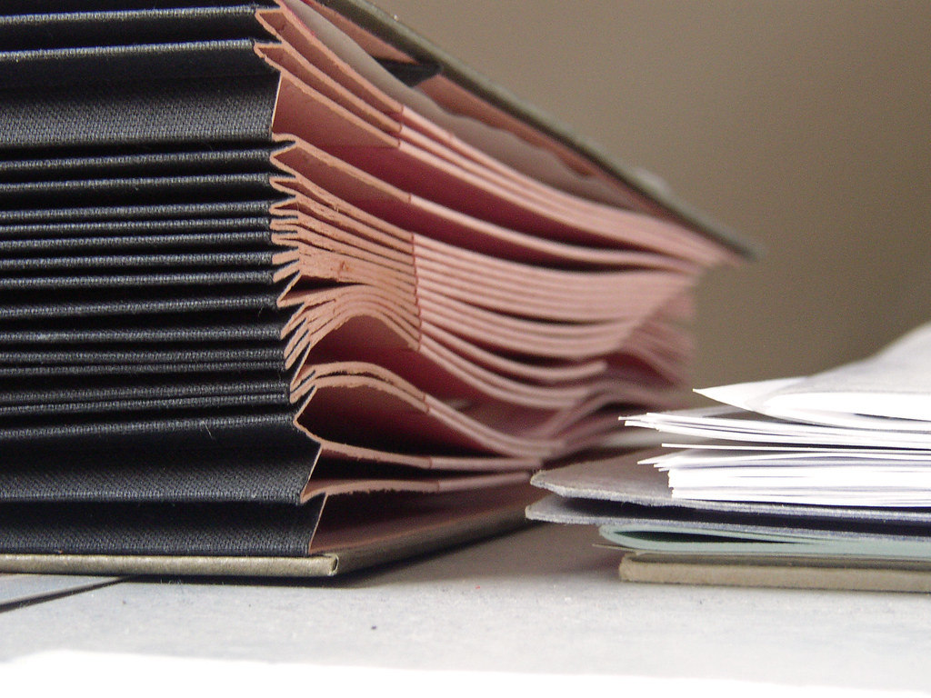 Close-up of a thick black book with fanned pink-edged pages beside a stack of loose white papers.