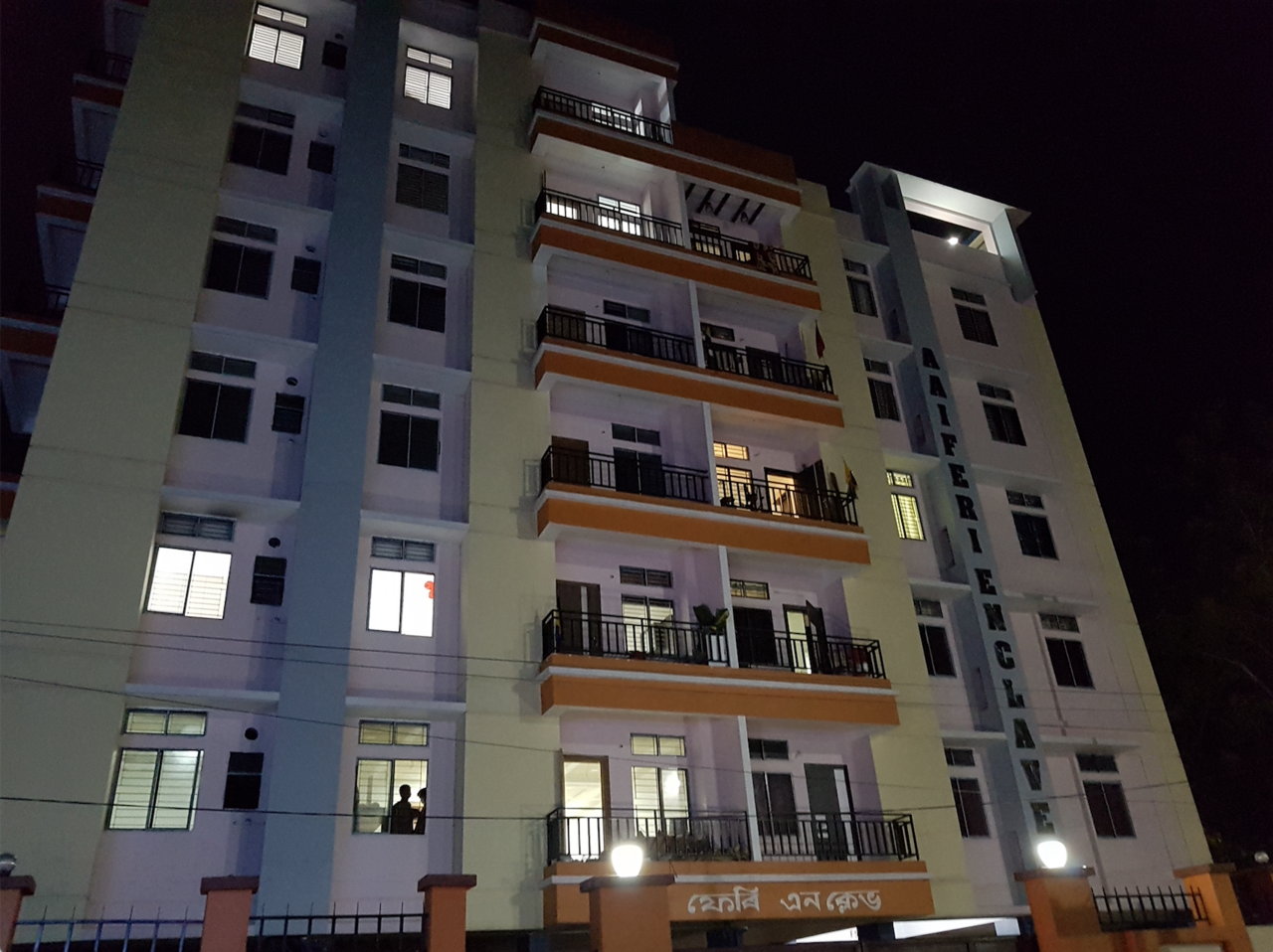 A multi-storey residential building at night with lit windows, balconies and a front boundary wall and gate.