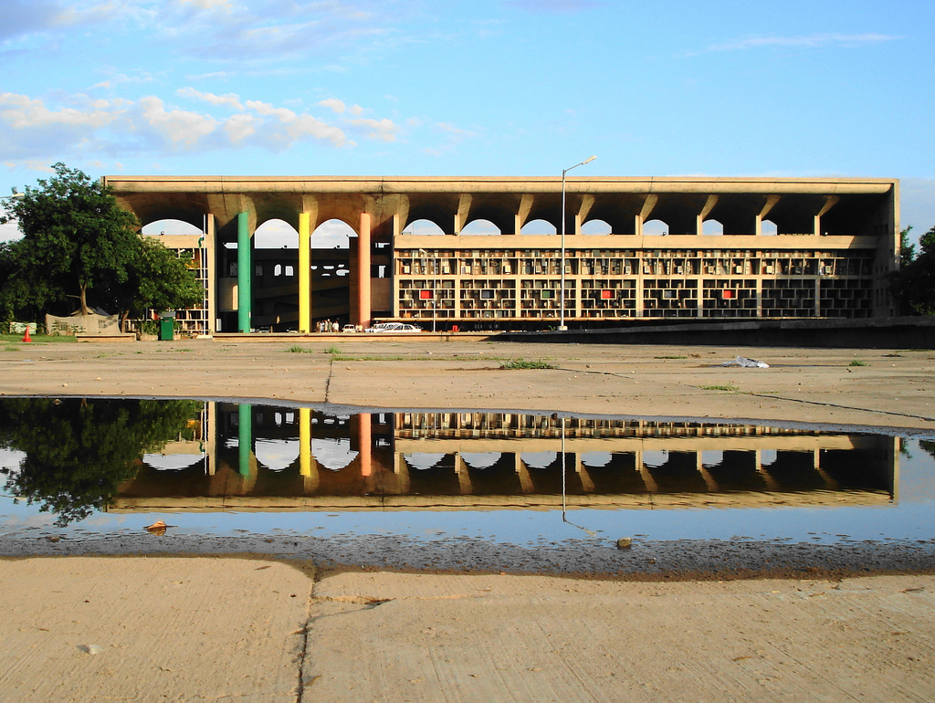 Modernist High Court building with coloured columns and arches, reflected in a large puddle on a concrete forecourt.