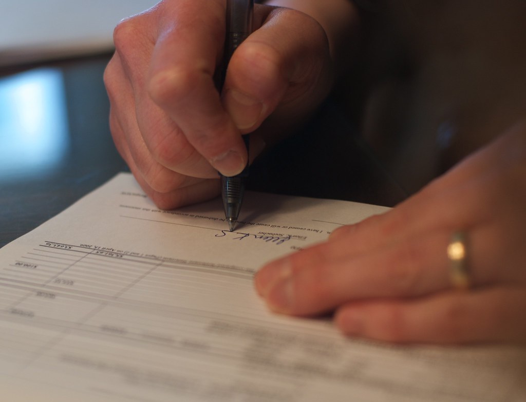 Close-up of hands signing a paper form with a black pen; a gold ring on one finger.