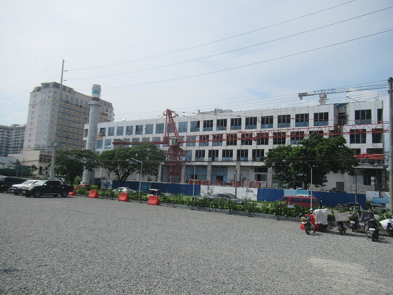 Construction site with a multi-storey building, red tower crane, red steel trusses, blue hoarding and parked vehicles