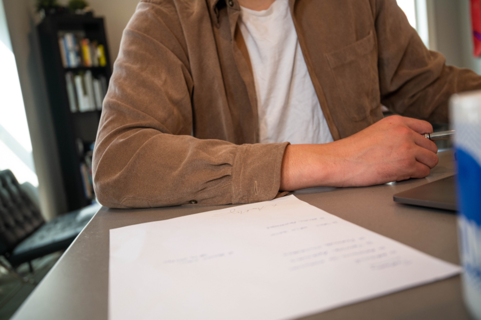 A person seated at an office desk with a printed sheet in front and a laptop to the side, holding a pen