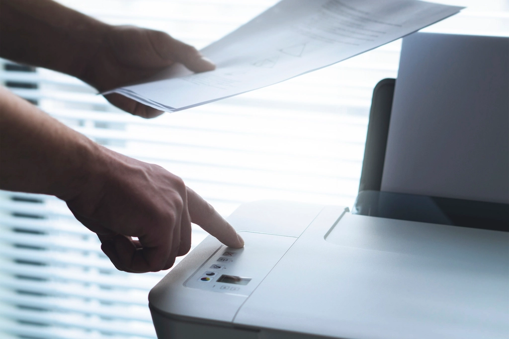 Close-up of hands holding sheets of paper above a printer while a finger presses a button on its control panel