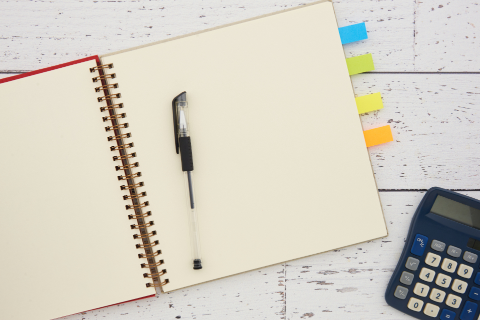 Open blank spiral notebook with a black pen, colored tabs and a calculator on a white wooden desk.