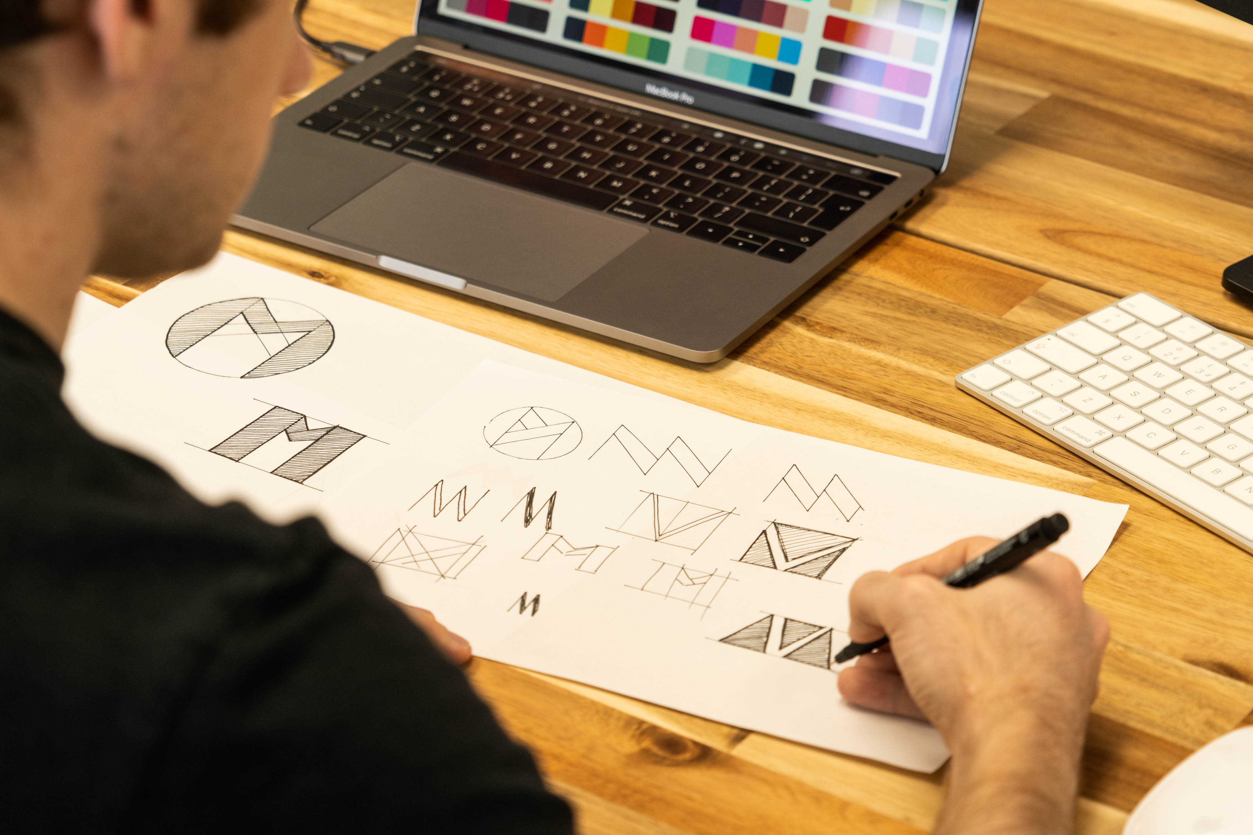 Hand sketching geometric logo ideas on paper beside a laptop showing a colour palette